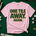 One Tile Away Again T-Shirt in Blossom, knotted flat lay on a green mahjong mat - "ONE TILE AWAY. AGAIN." in bold deep-green block letters with a gold drop shadow