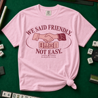 We Said Friendly Not Easy T-Shirt in Blossom, knotted flat lay on a green mahjong mat - a burgundy and pink club badge with a handshake over four mahjong tiles and the words "WE SAID FRIENDLY. NOT EASY. MAHJONG CLUB."