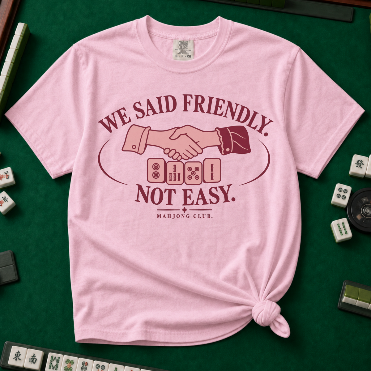 We Said Friendly Not Easy T-Shirt in Blossom, knotted flat lay on a green mahjong mat - a burgundy and pink club badge with a handshake over four mahjong tiles and the words "WE SAID FRIENDLY. NOT EASY. MAHJONG CLUB."