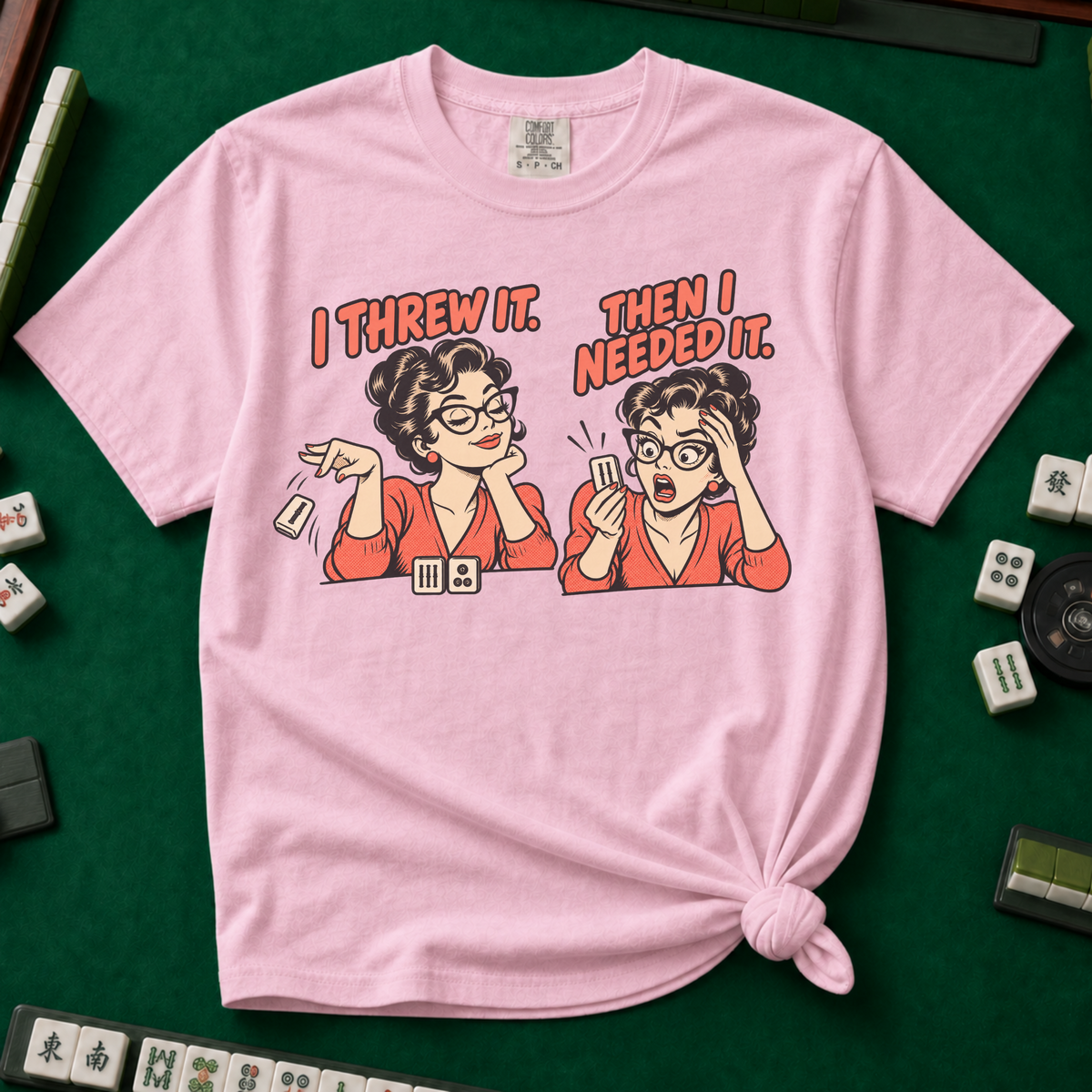 I Threw It Then I Needed It T-Shirt in Blossom, knotted flat lay on a green mahjong mat - a retro comic two-panel scene of a woman smugly flicking away a mahjong tile, then panicking when she needs it, with the words "I THREW IT. THEN I NEEDED IT."
