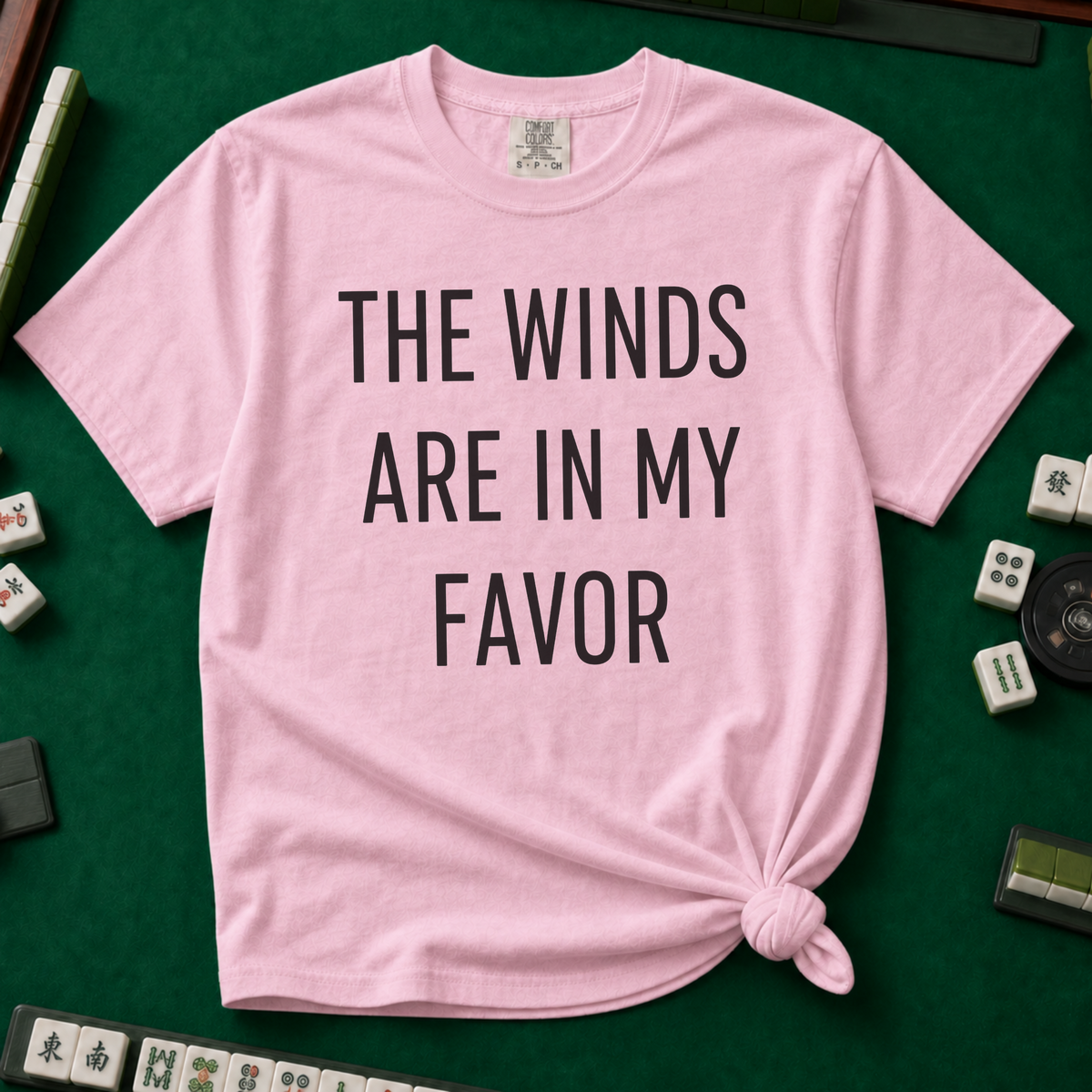 The Winds Are In My Favor T-Shirt in Blossom, knotted flat lay on a green mahjong mat - "THE WINDS ARE IN MY FAVOR" in tall black letters