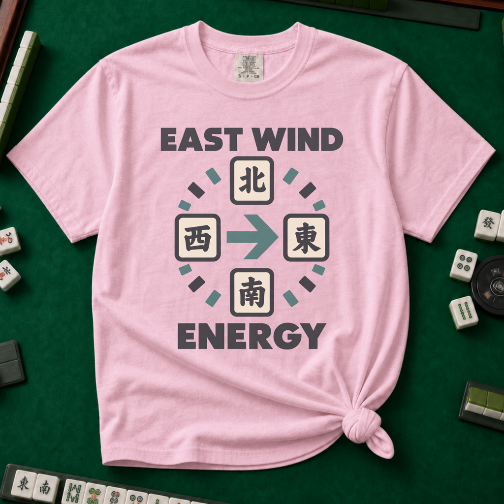 East Wind Energy T-Shirt in Blossom, knotted flat lay on a green mahjong mat - the four mahjong wind tiles arranged in a compass circle - North on top, West left, East right, South below - with a big teal arrow pointing East, between "EAST WIND" and "ENERGY" in charcoal block letters