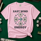 East Wind Energy T-Shirt in Blossom, knotted flat lay on a green mahjong mat - the four mahjong wind tiles arranged in a compass circle - North on top, West left, East right, South below - with a big teal arrow pointing East, between "EAST WIND" and "ENERGY" in charcoal block letters