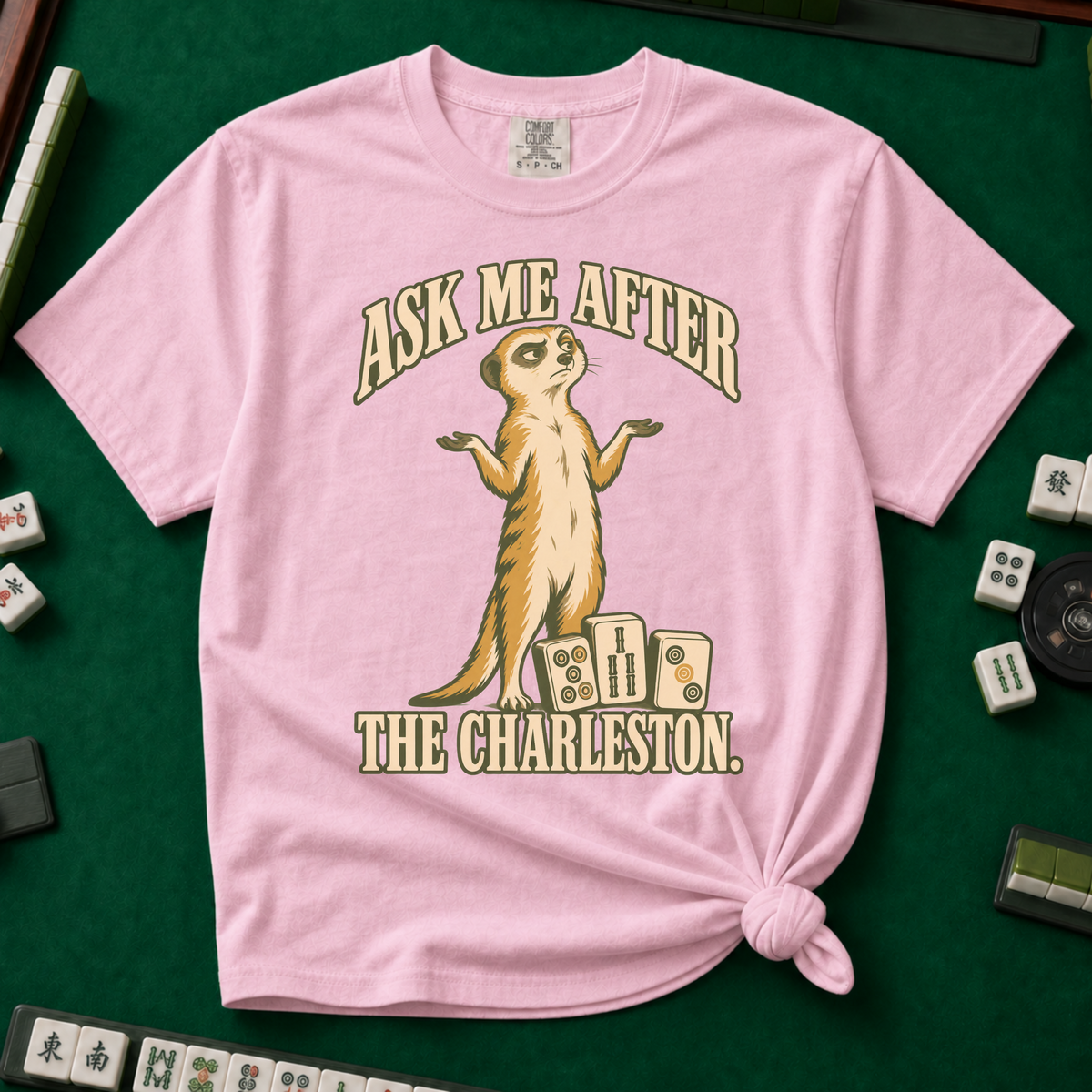Ask Me After The Charleston T-Shirt in Blossom, knotted flat lay on a green mahjong mat - shrugging meerkat standing beside three mahjong tiles - "ASK ME AFTER THE CHARLESTON." in cream collegiate letters