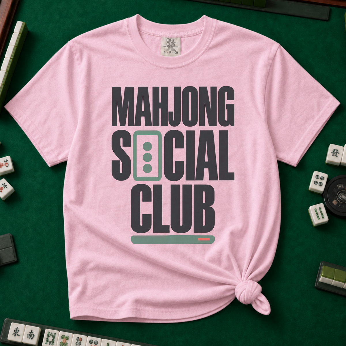 Mahjong Social Club Bold T-Shirt in Blossom, knotted flat lay on a green mahjong mat - bold charcoal block letters "MAHJONG SOCIAL CLUB" with a green three-dot tile as the O in SOCIAL and a green pusher bar underline