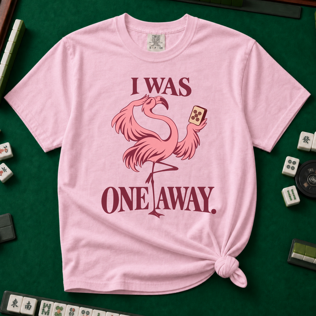 I Was One Away T-Shirt in Blossom - knotted tee flat lay on a green mahjong mat with tiles, printed with a pink flamingo standing on one leg and holding a mahjong tile, with the words I WAS ONE AWAY.