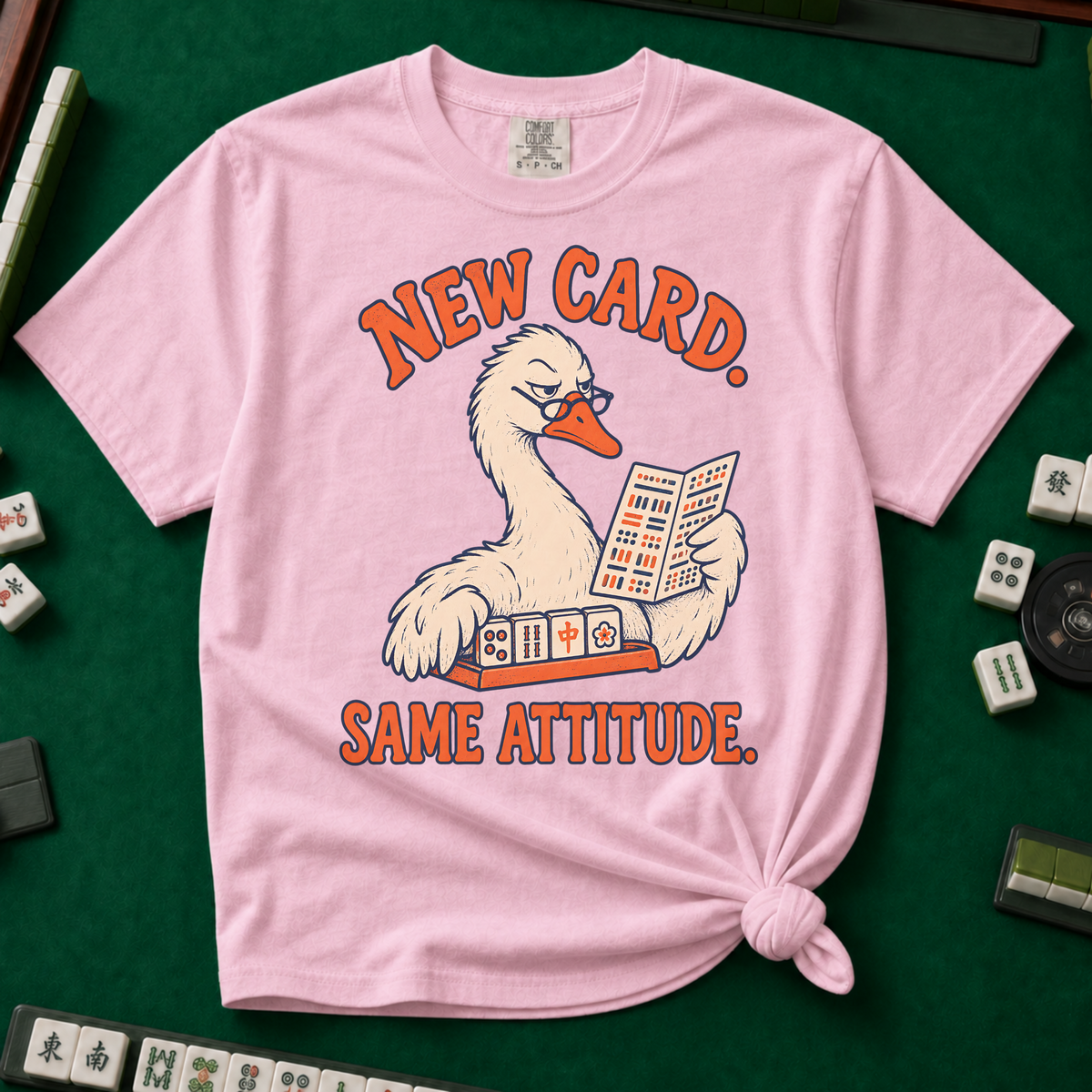 New Card T-Shirt in Blossom - knotted tee flat lay on a green mahjong mat with tiles, printed with a white goose in glasses reading the new mahjong card over a rack of tiles, with the words NEW CARD. SAME ATTITUDE.
