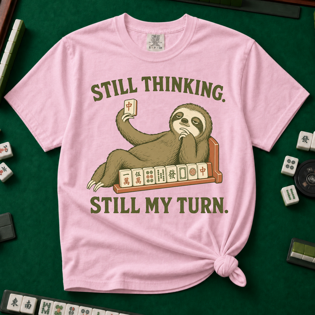 Still Thinking Still My Turn T-Shirt in Blossom, knotted flat lay on a green mahjong mat - a sloth draped along its mahjong tile rack, chin in hand, holding up a red dragon tile - "STILL THINKING. STILL MY TURN." in olive green vintage letters