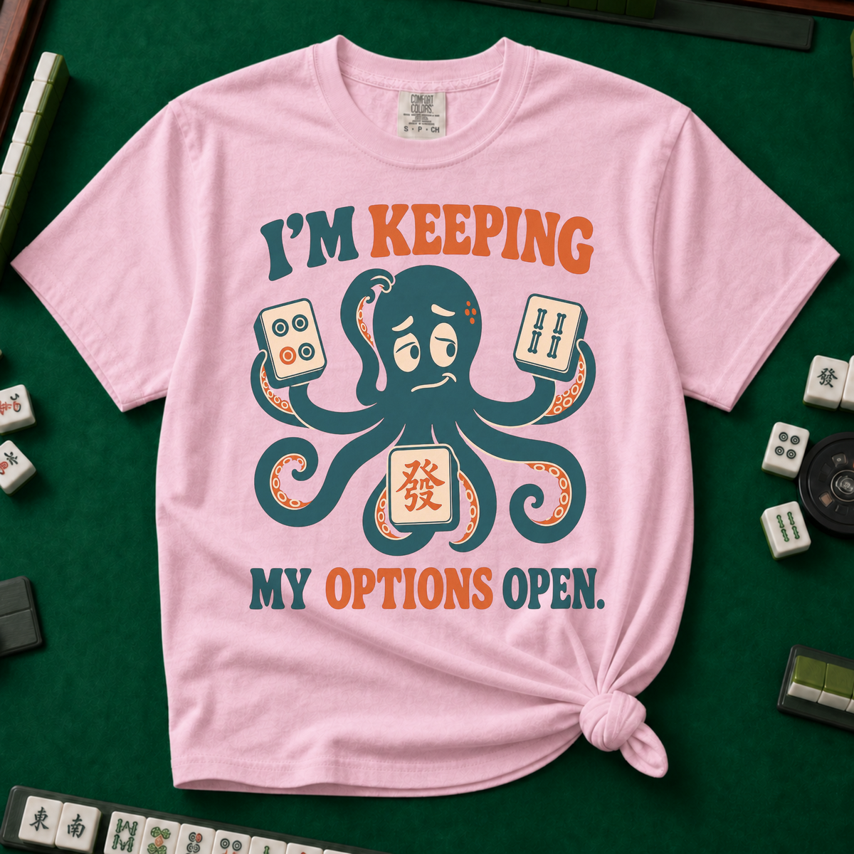 I'm Keeping My Options Open T-Shirt in Blossom, knotted flat lay on a green mahjong mat - worried teal octopus holding a dot tile, a bam tile, and a green dragon tile - "I'M KEEPING MY OPTIONS OPEN." in teal and orange retro letters