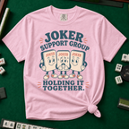 Joker Support Group Holding It Together T-Shirt in Blossom, knotted flat lay on a green mahjong mat - three retro cartoon joker tiles in sneakers, the middle one sweating nervously while the other two hold him up - "JOKER SUPPORT GROUP - HOLDING IT TOGETHER." in teal retro letters
