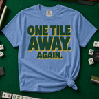 One Tile Away Again T-Shirt in Blue Jean, knotted flat lay on a green mahjong mat - "ONE TILE AWAY. AGAIN." in bold deep-green block letters with a gold drop shadow