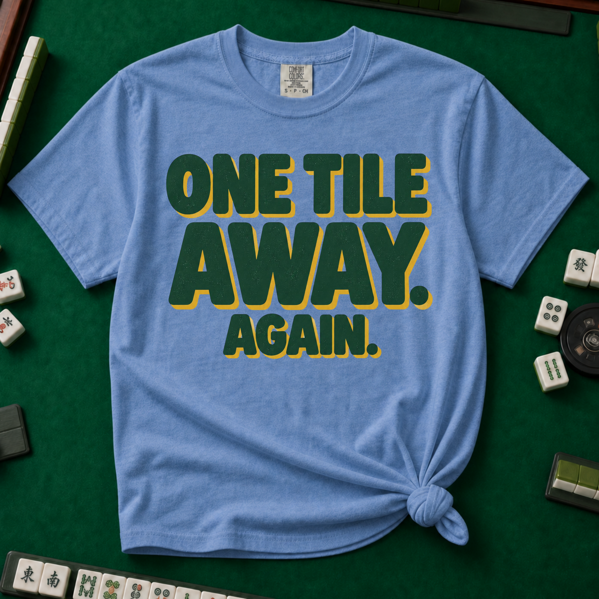 One Tile Away Again T-Shirt in Blue Jean, knotted flat lay on a green mahjong mat - "ONE TILE AWAY. AGAIN." in bold deep-green block letters with a gold drop shadow
