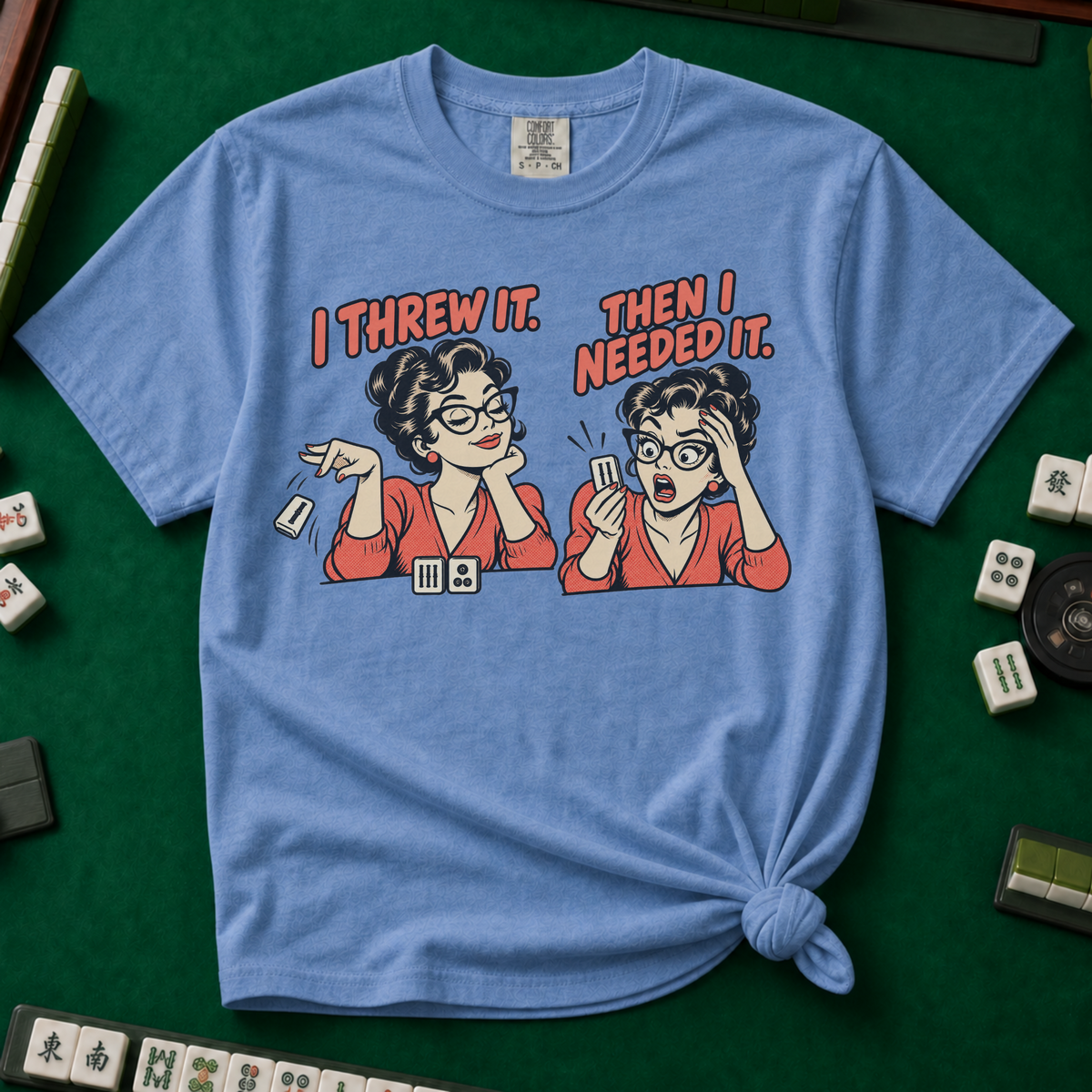 I Threw It Then I Needed It T-Shirt in Blue Jean, knotted flat lay on a green mahjong mat - a retro comic two-panel scene of a woman smugly flicking away a mahjong tile, then panicking when she needs it, with the words "I THREW IT. THEN I NEEDED IT."