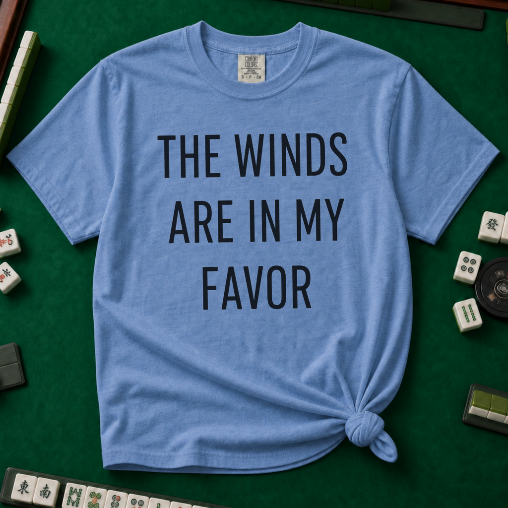 The Winds Are In My Favor T-Shirt in Blue Jean, knotted flat lay on a green mahjong mat - "THE WINDS ARE IN MY FAVOR" in tall black letters