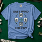 East Wind Energy T-Shirt in Blue Jean, knotted flat lay on a green mahjong mat - the four mahjong wind tiles arranged in a compass circle - North on top, West left, East right, South below - with a big teal arrow pointing East, between "EAST WIND" and "ENERGY" in charcoal block letters
