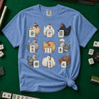 Mahjong Chickens T-Shirt in Blue Jean, knotted flat lay on a green mahjong mat - a 3x3 grid of nine hens, each posing with a mahjong tile - a sunglasses hen with the green dragon, a white hen with the red dragon, a black hen with dots, a crested hen with East, and more