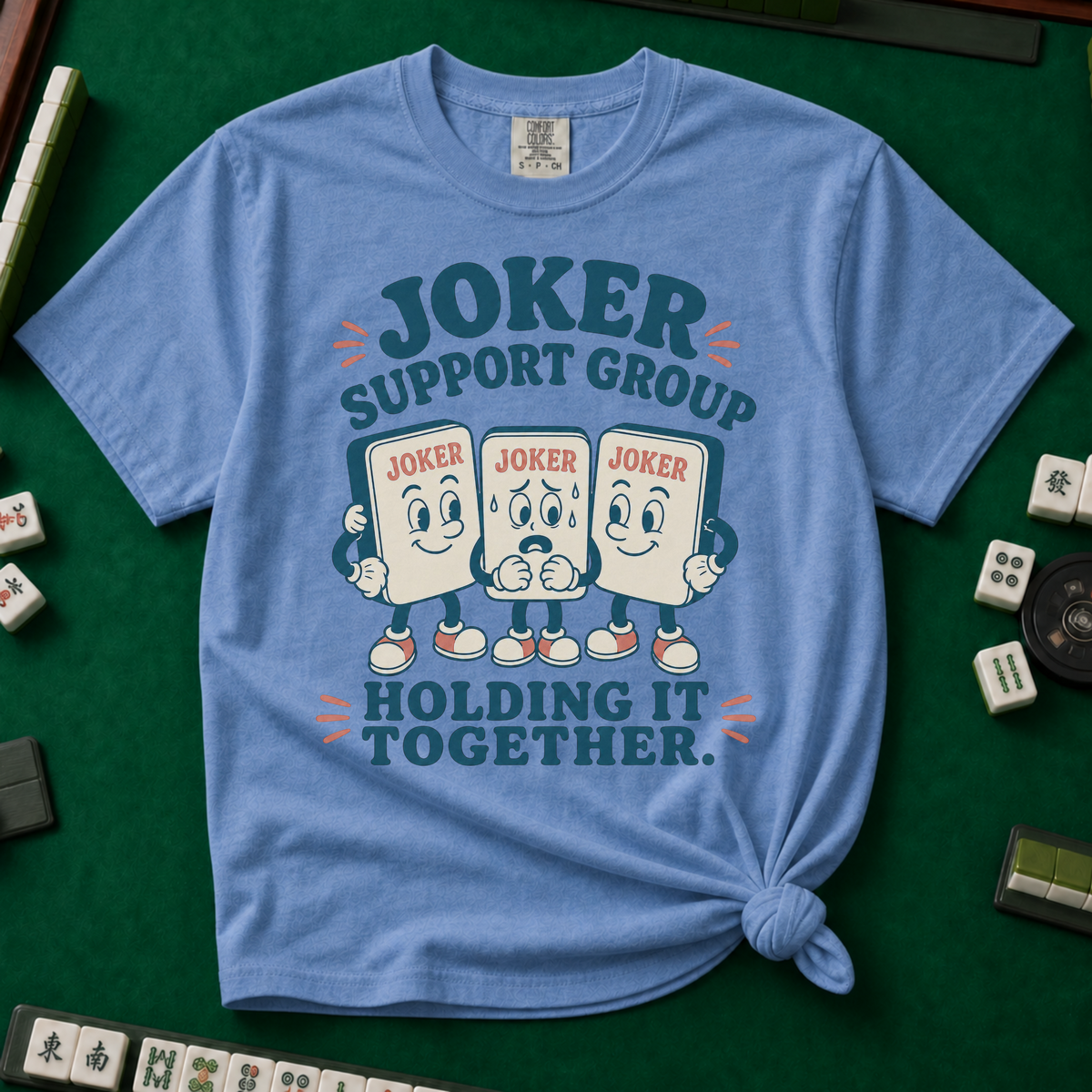 Joker Support Group Holding It Together T-Shirt in Blue Jean, knotted flat lay on a green mahjong mat - three retro cartoon joker tiles in sneakers, the middle one sweating nervously while the other two hold him up - "JOKER SUPPORT GROUP - HOLDING IT TOGETHER." in teal retro letters