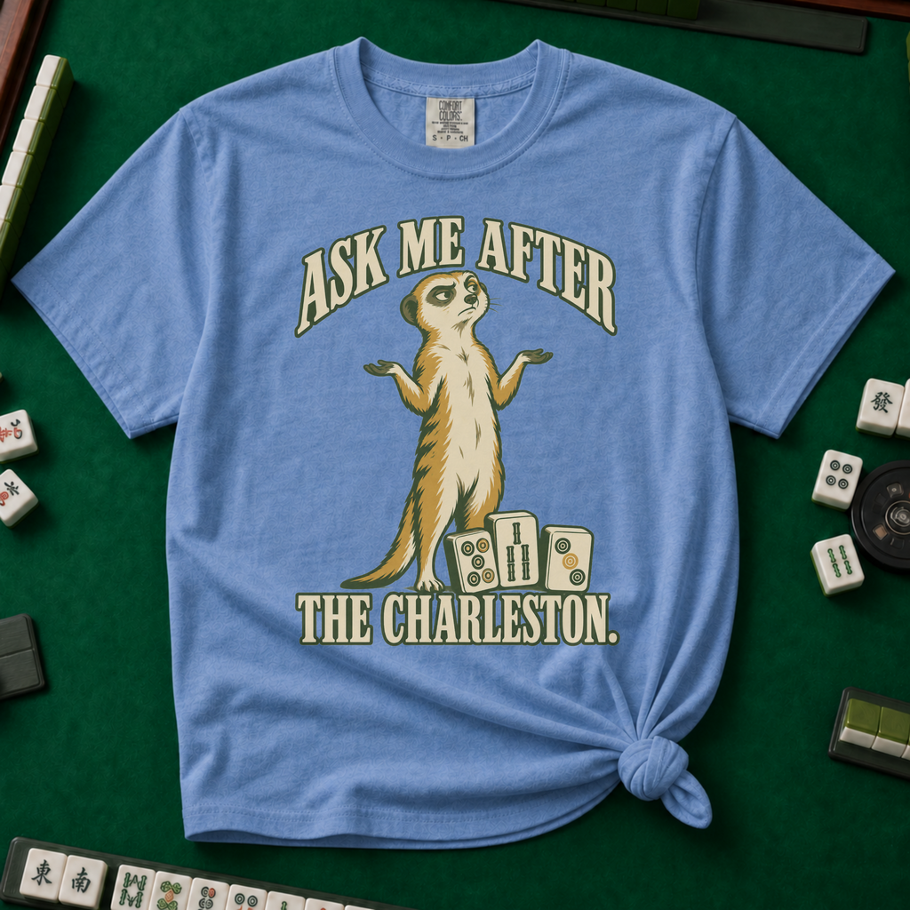Ask Me After The Charleston T-Shirt in Blue Jean, knotted flat lay on a green mahjong mat - shrugging meerkat standing beside three mahjong tiles - "ASK ME AFTER THE CHARLESTON." in cream collegiate letters