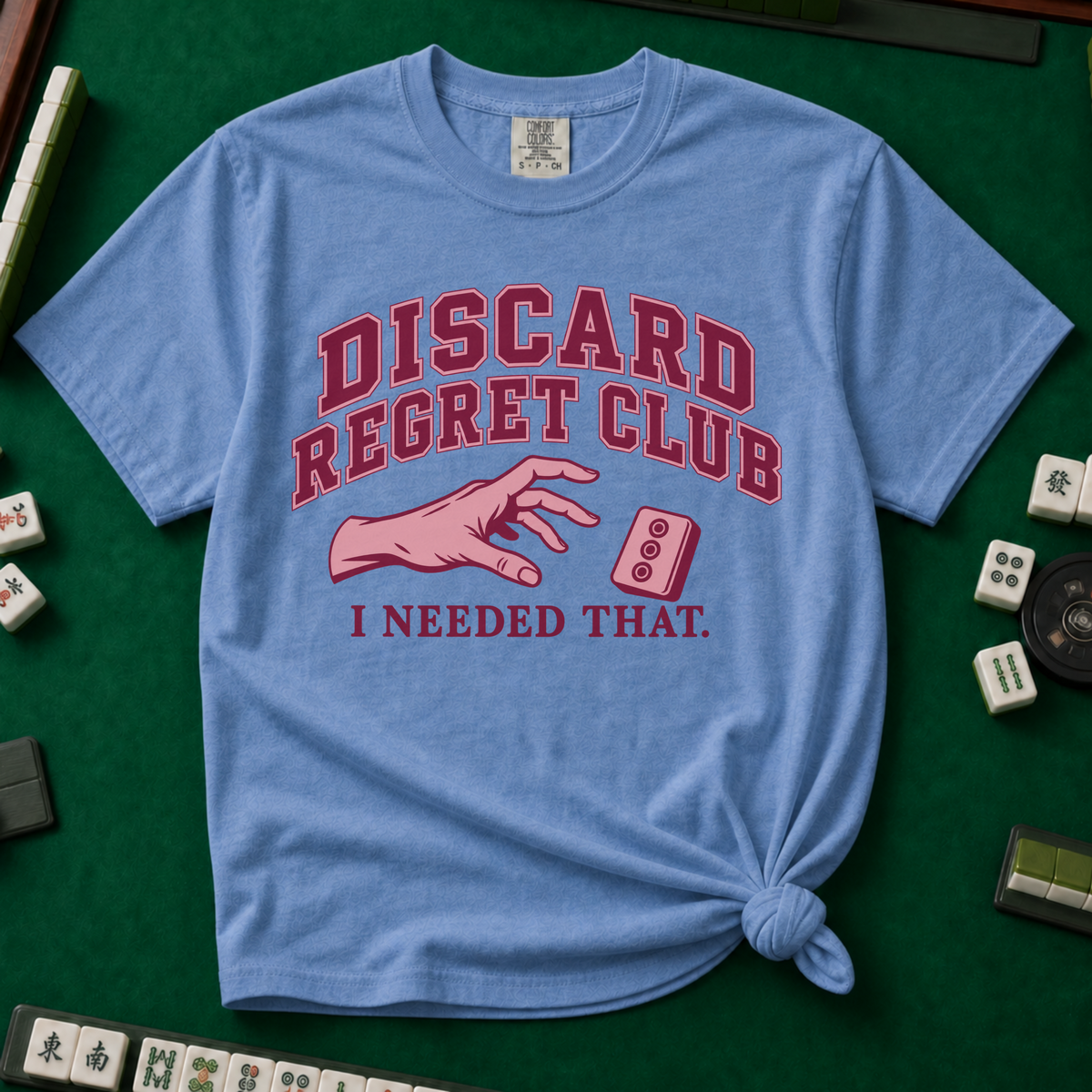 Discard Regret Club I Needed That T-Shirt in Blue Jean, knotted flat lay on a green mahjong mat - burgundy varsity letters "DISCARD REGRET CLUB" over a pink hand reaching back for a pink dot tile it just discarded - "I NEEDED THAT."