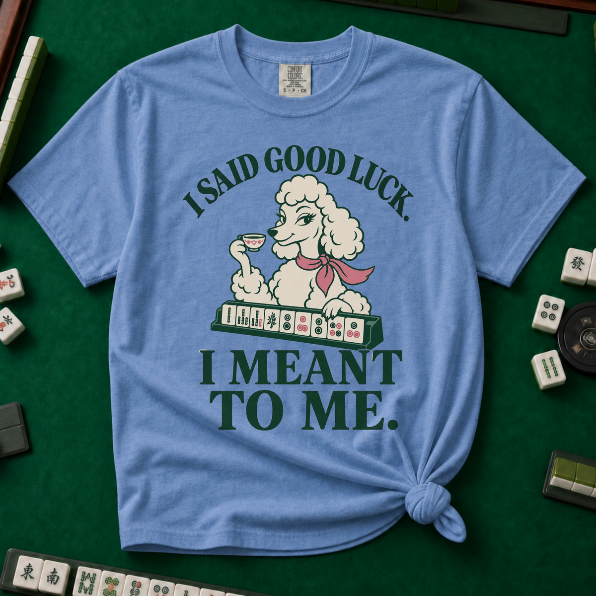 I Said Good Luck T-Shirt in Blue Jean - knotted tee flat lay on a green mahjong mat with tiles, printed with a cream poodle in a pink scarf holding a teacup behind a rack of mahjong tiles, with the words I SAID GOOD LUCK. I MEANT TO ME.