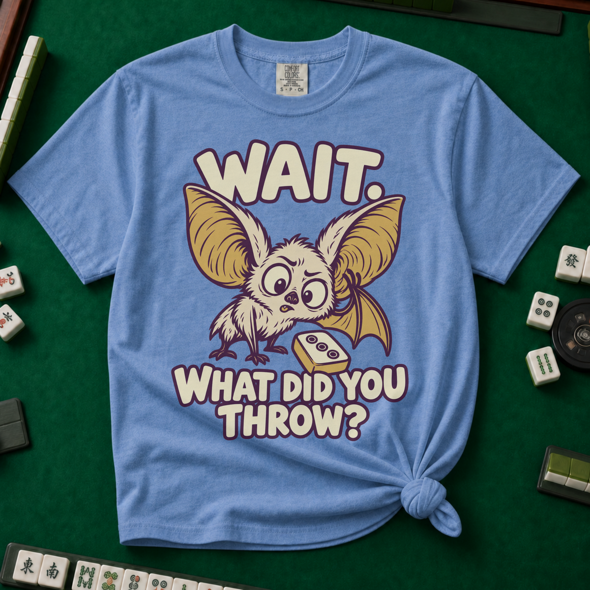 Wait What Did You Throw T-Shirt in Blue Jean, knotted flat lay on a green mahjong mat - a wide-eyed fluffy bat with huge golden ears leaning in over a fallen dot tile - "WAIT. WHAT DID YOU THROW?" in cream bubble letters