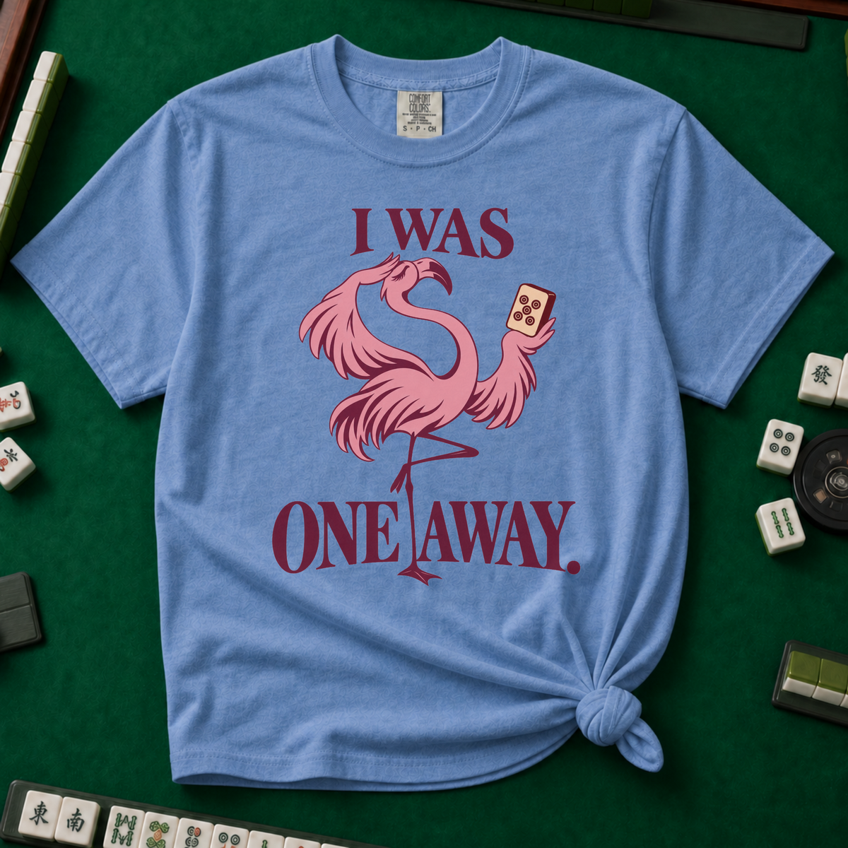 I Was One Away T-Shirt in Blue Jean - knotted tee flat lay on a green mahjong mat with tiles, printed with a pink flamingo standing on one leg and holding a mahjong tile, with the words I WAS ONE AWAY.