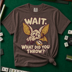 Wait What Did You Throw T-Shirt in Espresso, knotted flat lay on a green mahjong mat - a wide-eyed fluffy bat with huge golden ears leaning in over a fallen dot tile - "WAIT. WHAT DID YOU THROW?" in cream bubble letters