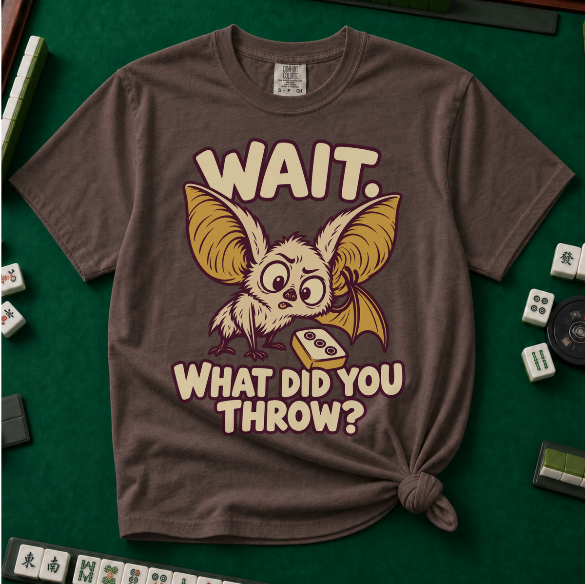 Wait What Did You Throw T-Shirt in Espresso, knotted flat lay on a green mahjong mat - a wide-eyed fluffy bat with huge golden ears leaning in over a fallen dot tile - "WAIT. WHAT DID YOU THROW?" in cream bubble letters