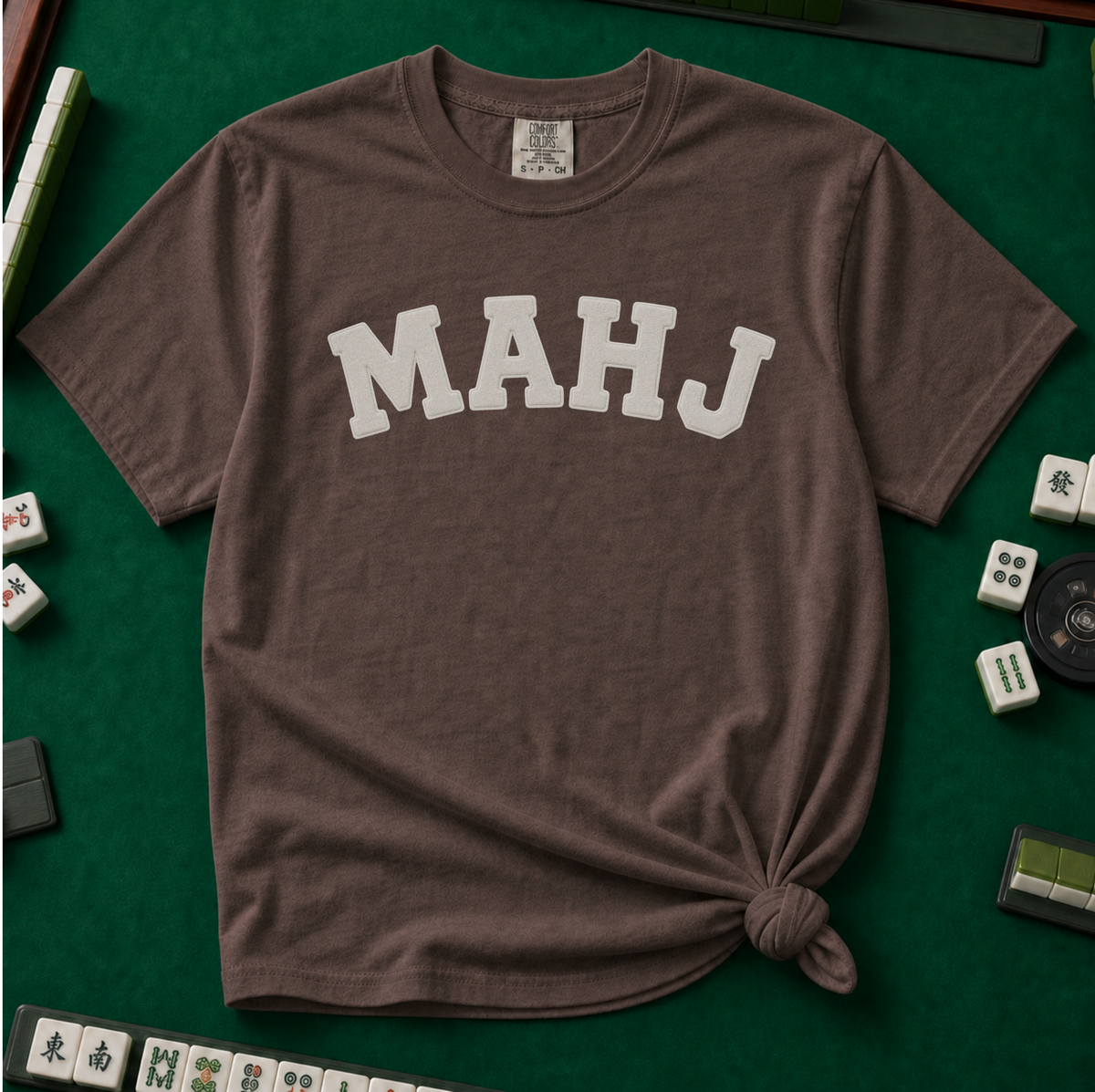 MAHJ Varsity T-Shirt in Espresso, knotted flat lay on a green mahjong mat - "MAHJ" in big white varsity arch letters with a stitched letterman look