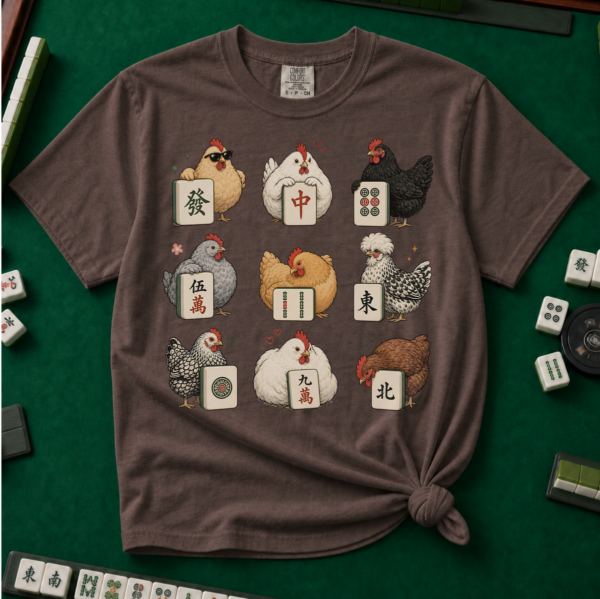 Mahjong Chickens T-Shirt in Espresso, knotted flat lay on a green mahjong mat - a 3x3 grid of nine hens, each posing with a mahjong tile - a sunglasses hen with the green dragon, a white hen with the red dragon, a black hen with dots, a crested hen with East, and more