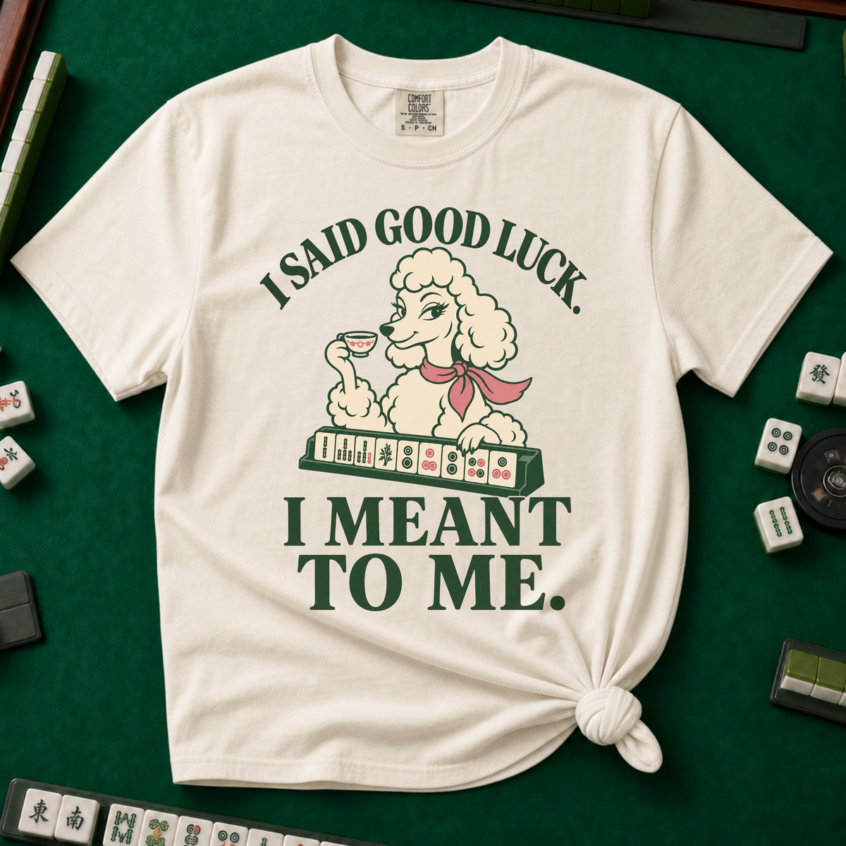 I Said Good Luck T-Shirt in Ivory - knotted tee flat lay on a green mahjong mat with tiles, printed with a cream poodle in a pink scarf holding a teacup behind a rack of mahjong tiles, with the words I SAID GOOD LUCK. I MEANT TO ME.