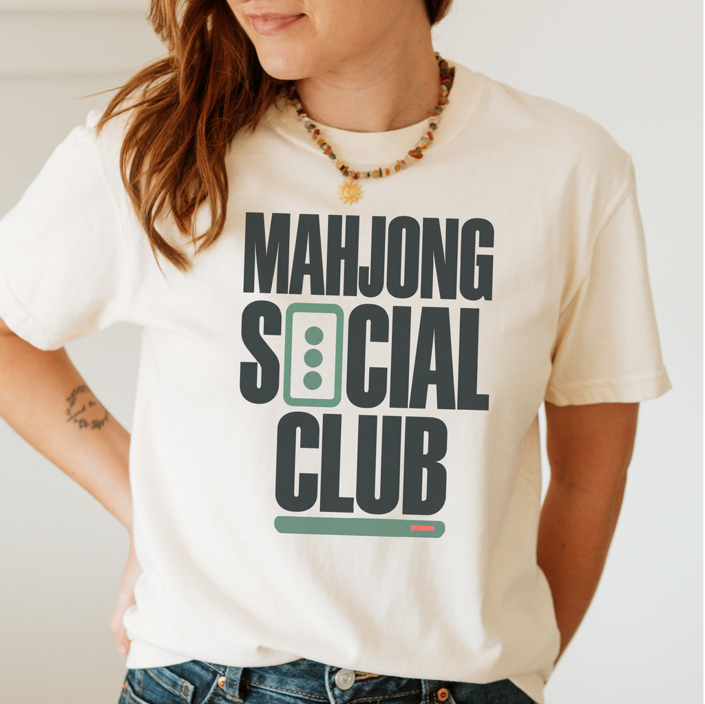 Woman wearing the Mahjong Social Club Bold T-Shirt in Ivory - bold charcoal block letters "MAHJONG SOCIAL CLUB" with a green three-dot tile as the O in SOCIAL and a green pusher bar underline
