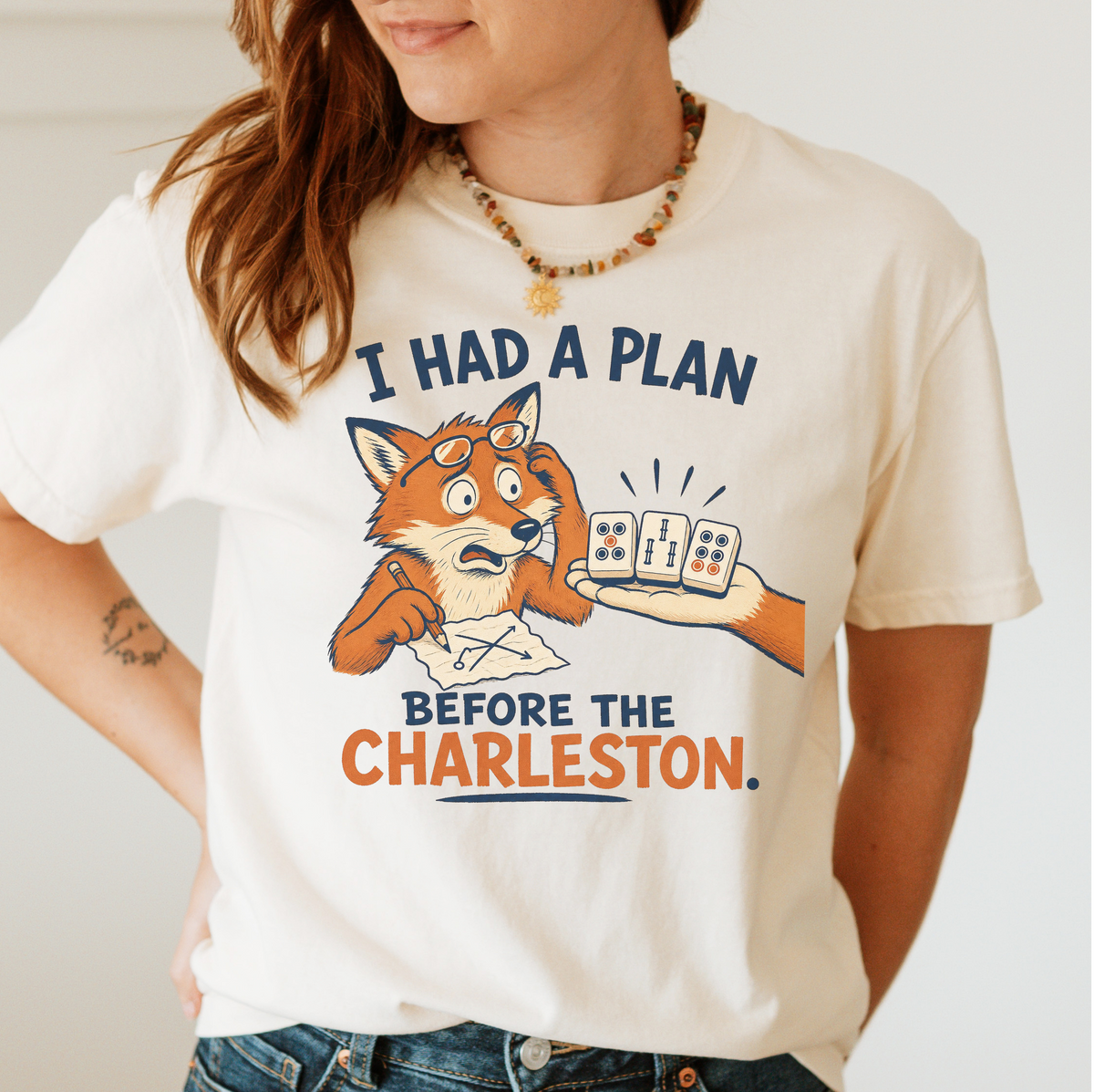 Woman wearing the I Had A Plan Before The Charleston T-Shirt in Ivory - a stressed cartoon fox with glasses and a strategy sketch being handed three mahjong tiles, with the words "I HAD A PLAN BEFORE THE CHARLESTON."