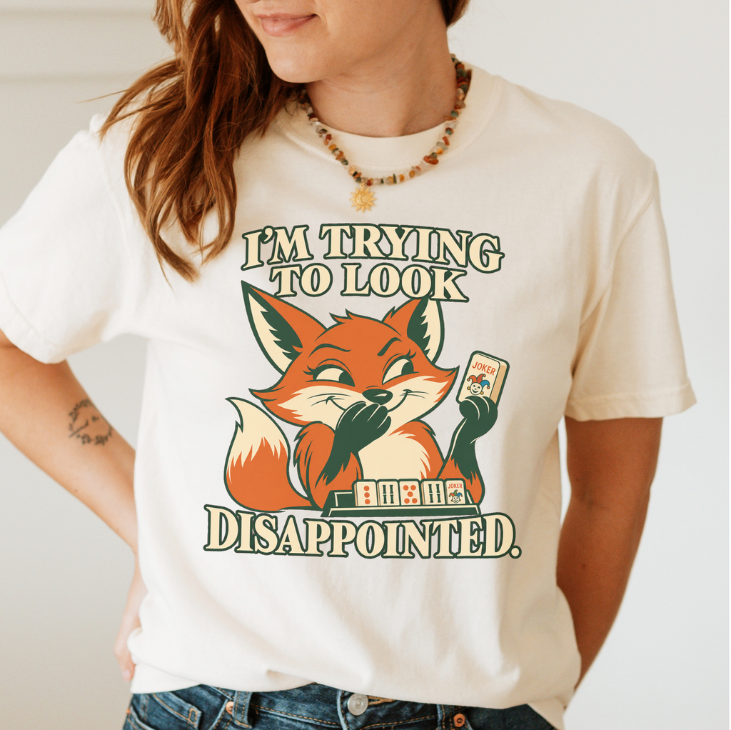 Woman wearing the I'm Trying To Look Disappointed T-Shirt in Ivory - smug retro fox holding up a joker tile, hiding a grin behind one paw, tile rack in front - "I'M TRYING TO LOOK DISAPPOINTED." in cream collegiate letters