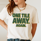 Woman wearing the One Tile Away Again T-Shirt in Ivory - "ONE TILE AWAY. AGAIN." in bold deep-green block letters with a gold drop shadow