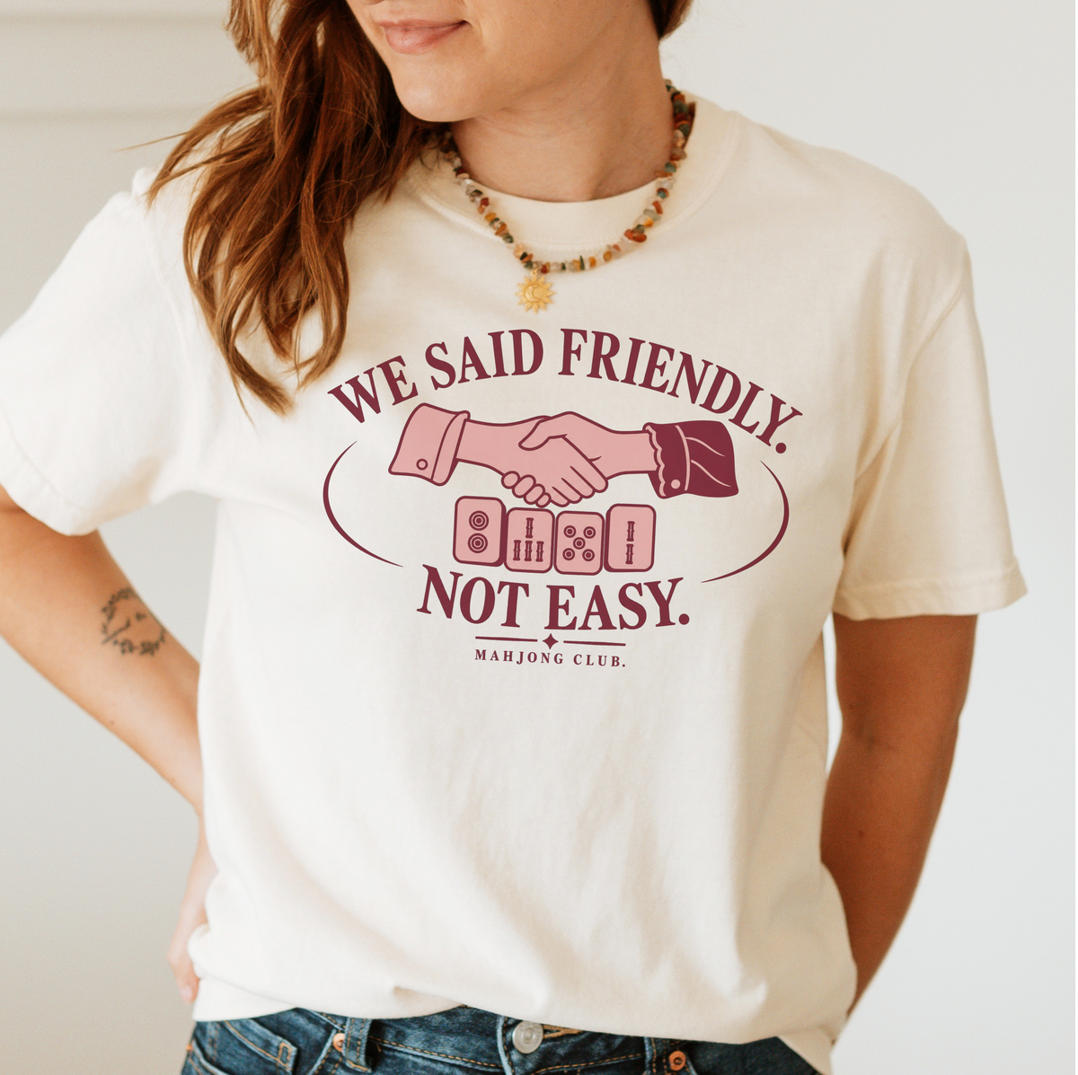 Woman wearing the We Said Friendly Not Easy T-Shirt in Ivory - a burgundy and pink club badge with a handshake over four mahjong tiles and the words "WE SAID FRIENDLY. NOT EASY. MAHJONG CLUB."