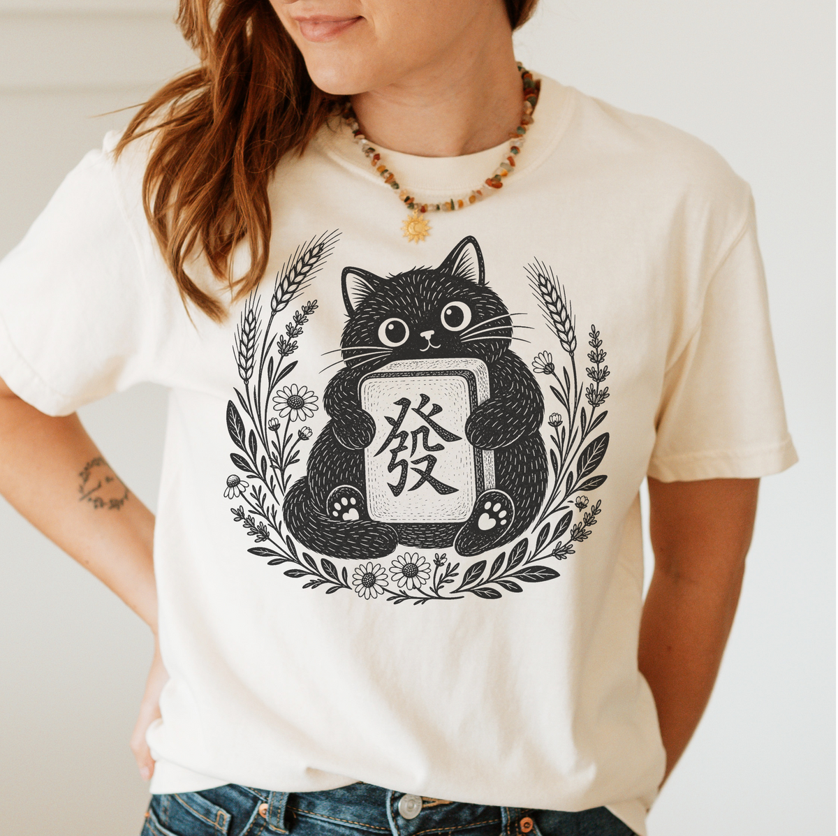 Woman wearing the Linocut Tile Cat T-Shirt in Ivory - a linocut-style black cat hugging a Green Dragon mahjong tile inside a wreath of wheat, daisies and lavender