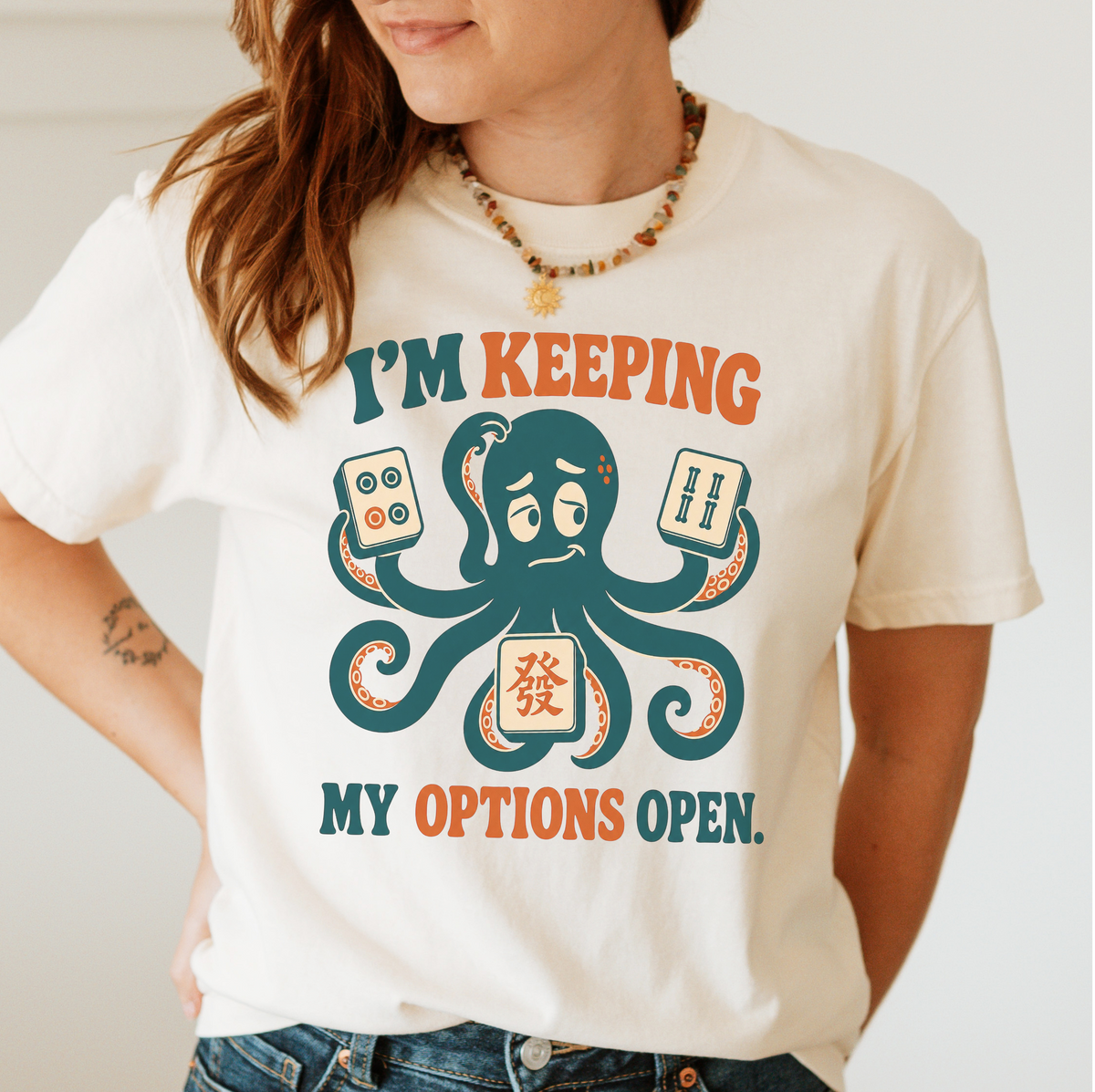 Woman wearing the I'm Keeping My Options Open T-Shirt in Ivory - worried teal octopus holding a dot tile, a bam tile, and a green dragon tile - "I'M KEEPING MY OPTIONS OPEN." in teal and orange retro letters
