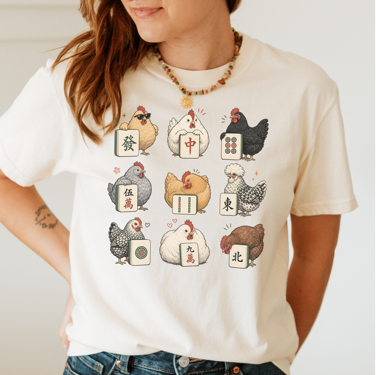 Woman wearing the Mahjong Chickens T-Shirt in Ivory - a 3x3 grid of nine hens, each posing with a mahjong tile - a sunglasses hen with the green dragon, a white hen with the red dragon, a black hen with dots, a crested hen with East, and more