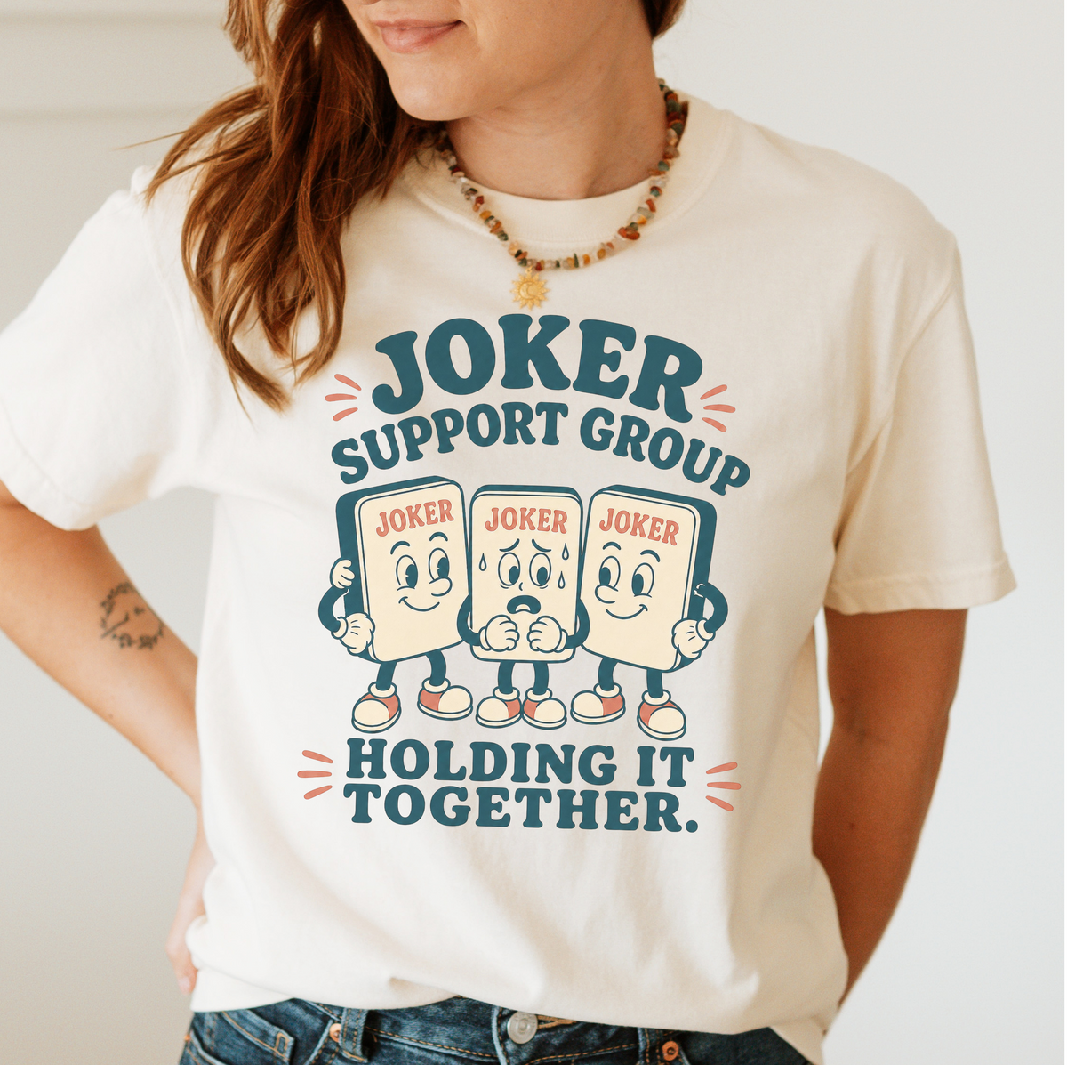 Woman wearing the Joker Support Group Holding It Together T-Shirt in Ivory - three retro cartoon joker tiles in sneakers, the middle one sweating nervously while the other two hold him up - "JOKER SUPPORT GROUP - HOLDING IT TOGETHER." in teal retro letters