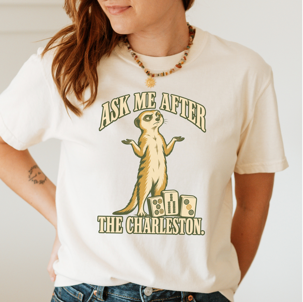 Woman wearing the Ask Me After The Charleston T-Shirt in Ivory - shrugging meerkat standing beside three mahjong tiles - "ASK ME AFTER THE CHARLESTON." in cream collegiate letters