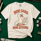 New Card T-Shirt in Ivory - knotted tee flat lay on a green mahjong mat with tiles, printed with a white goose in glasses reading the new mahjong card over a rack of tiles, with the words NEW CARD. SAME ATTITUDE.