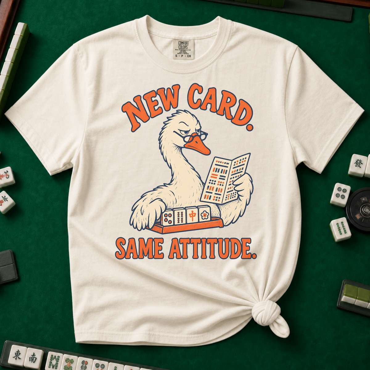 New Card T-Shirt in Ivory - knotted tee flat lay on a green mahjong mat with tiles, printed with a white goose in glasses reading the new mahjong card over a rack of tiles, with the words NEW CARD. SAME ATTITUDE.