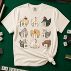Mahjong Chickens T-Shirt in Ivory, knotted flat lay on a green mahjong mat - a 3x3 grid of nine hens, each posing with a mahjong tile - a sunglasses hen with the green dragon, a white hen with the red dragon, a black hen with dots, a crested hen with East, and more