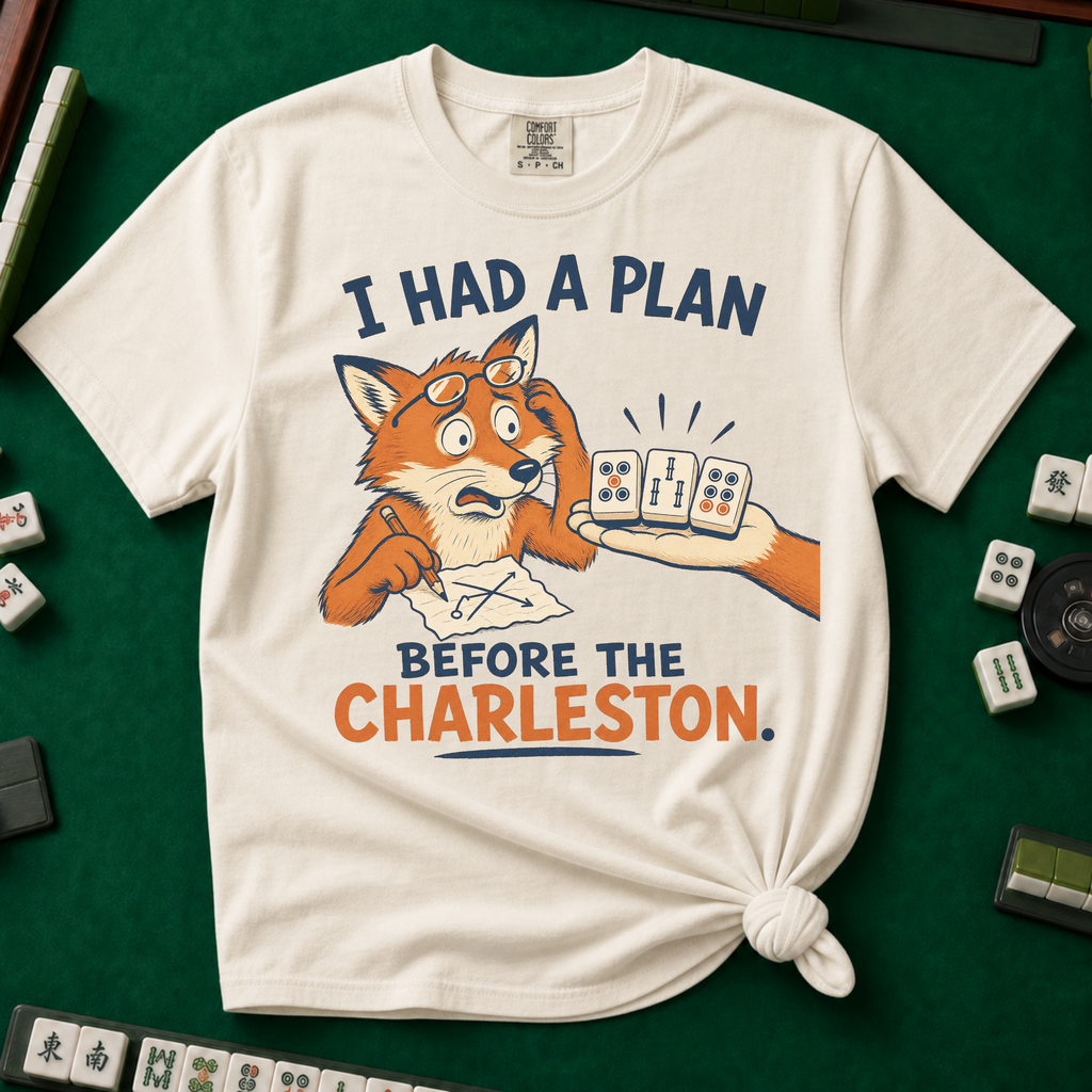 I Had A Plan Before The Charleston T-Shirt in Ivory, knotted flat lay on a green mahjong mat - a stressed cartoon fox with glasses and a strategy sketch being handed three mahjong tiles, with the words "I HAD A PLAN BEFORE THE CHARLESTON."