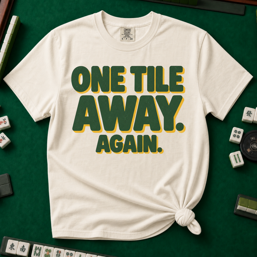 One Tile Away Again T-Shirt in Ivory, knotted flat lay on a green mahjong mat - "ONE TILE AWAY. AGAIN." in bold deep-green block letters with a gold drop shadow