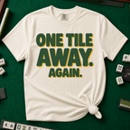 One Tile Away Again T-Shirt in Ivory, knotted flat lay on a green mahjong mat - "ONE TILE AWAY. AGAIN." in bold deep-green block letters with a gold drop shadow