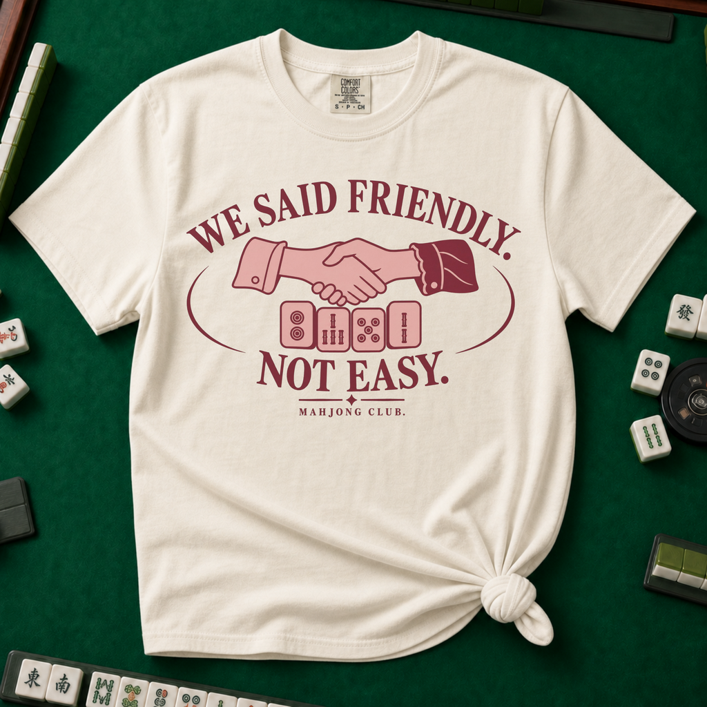 We Said Friendly Not Easy T-Shirt in Ivory, knotted flat lay on a green mahjong mat - a burgundy and pink club badge with a handshake over four mahjong tiles and the words "WE SAID FRIENDLY. NOT EASY. MAHJONG CLUB."