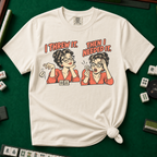 I Threw It Then I Needed It T-Shirt in Ivory, knotted flat lay on a green mahjong mat - a retro comic two-panel scene of a woman smugly flicking away a mahjong tile, then panicking when she needs it, with the words "I THREW IT. THEN I NEEDED IT."