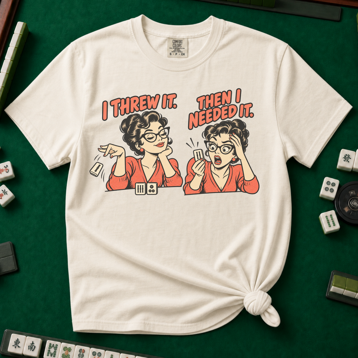 I Threw It Then I Needed It T-Shirt in Ivory, knotted flat lay on a green mahjong mat - a retro comic two-panel scene of a woman smugly flicking away a mahjong tile, then panicking when she needs it, with the words "I THREW IT. THEN I NEEDED IT."